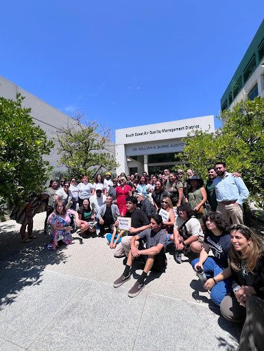 Dozens of smiling activists pose for a photo outside of South Coast Air Quality Management District Headquarters, two activists are holding signs which read “no more deaths” and “clean air now.” They are surrounded by trees and the sky is bright blue.