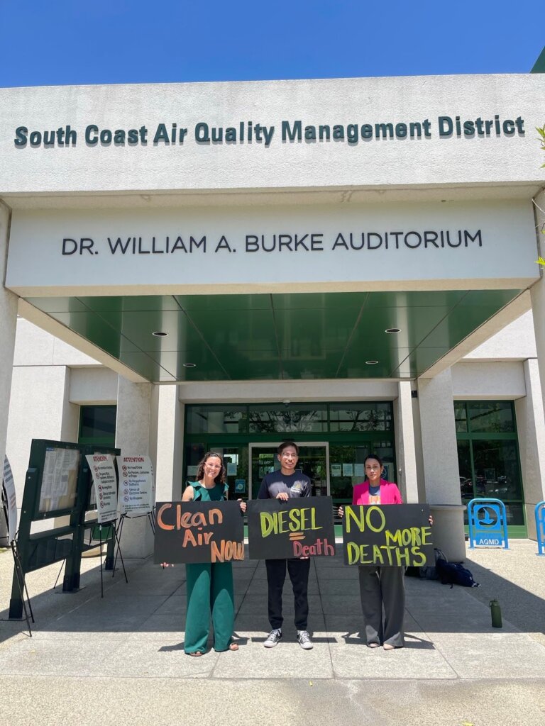 Three smiling advocates holding signs in favor of a strong Railyard ISR stand in front of the Dr. William A. Burke Auditorium at South Coast Air Quality Management District Headquarters. The signs read from left to right: “clean air now,” “diesel = death” and “no more deaths.”